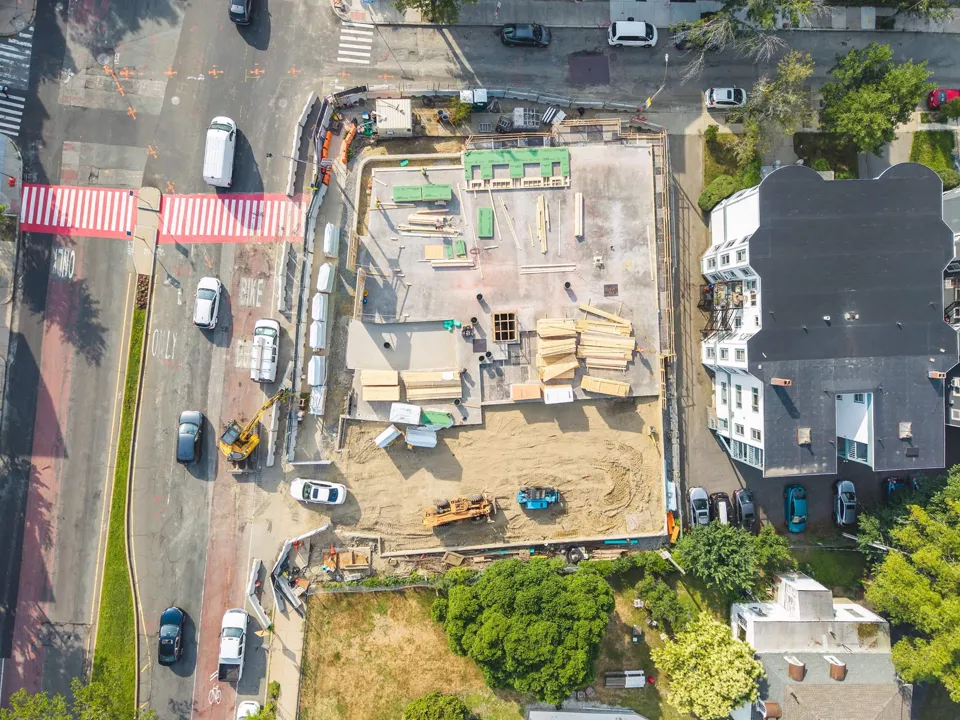 Overhead view of an organized multifamily jobsite with excavation, staged materials and a neighboring triple-decker